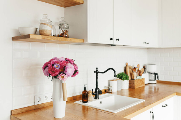 Uitzicht op schoon wit eenvoudige moderne keuken in Scandinavische stijl, keuken details, houten tafel, aster Michaelmas madeliefje boeket in vaas op de tafel - Foto, afbeelding