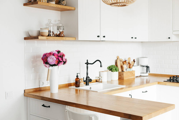 Blick auf sauberes Weiß einfache moderne Küche im skandinavischen Stil, Küchendetails, Holztisch, Aster Michaelmas Gänseblümchenstrauß in Vase auf dem Tisch - Foto, Bild