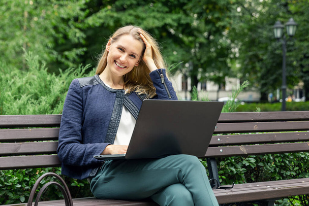 jonge aantrekkelijke glimlachende vrouw ontspannen op de bank in het stadspark met laptop - Foto, afbeelding