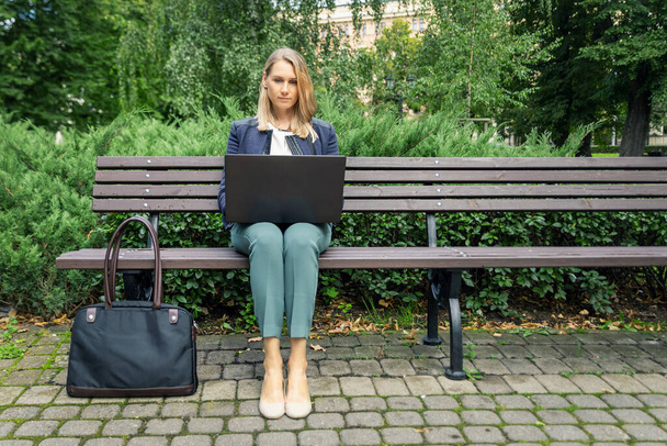 μια νεαρή επιχειρηματίας που κάθεται στον πάγκο στο πάρκο της πόλης και δουλεύει σε λάπτοπ. εξωτερική ανεξάρτητη εργασία γραφείου - Φωτογραφία, εικόνα
