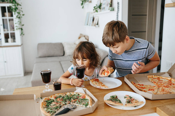 Yeme konseptinin keyfini çıkarın. Çocuklar evde pizza yiyorlar. İki sevimli çocuk çok mutlu duygusal yüz ifadeleri ve duruş ifadeleriyle pizza yemekten hoşlanırlar.. - Fotoğraf, Görsel
