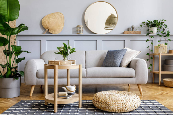 Scandinavian living room interior with design grey sofa, wooden coffee table, tropical plants, shelf, mirror, furniture, plaid pillow, teapot, book and elegant personal accessories in home decor. - Foto, Imagen