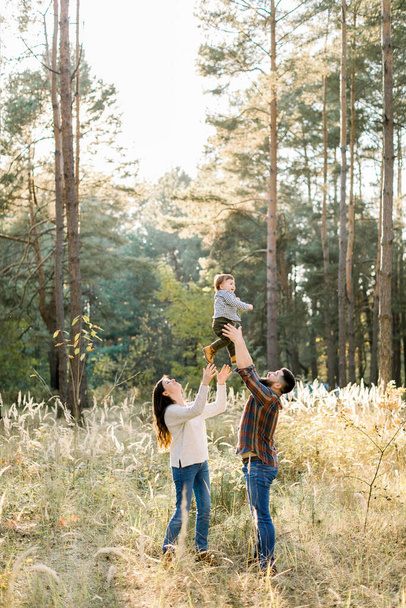 Outdoor portrait of happy young parents, having fun and lifting up their little cute baby son, during walk in autumn forest at sunny day - 写真・画像