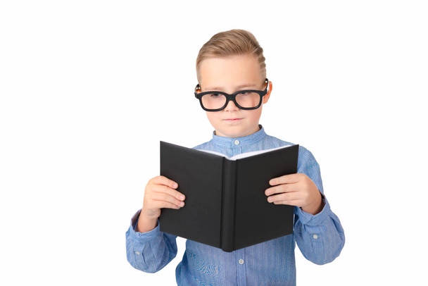 Mignon écolier sérieux lit livre et porter des lunettes tout en étant debout dans le studio, isolé sur blanc - Photo, image