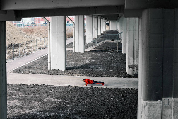 Young attractive sportsman in tracksuit and red sweatshirt doing push-ups in urban exterior. - Foto, Imagen