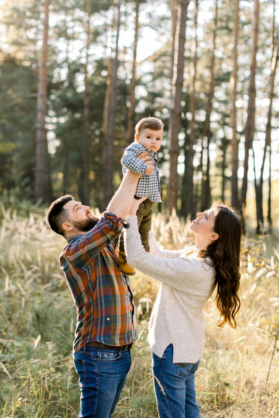 Outdoor family portrait of happy young parents, wearing stylish casual clothes, having fun and lifting up their little cute baby son, during walk in autumn forest at sunny day - Zdjęcie, obraz