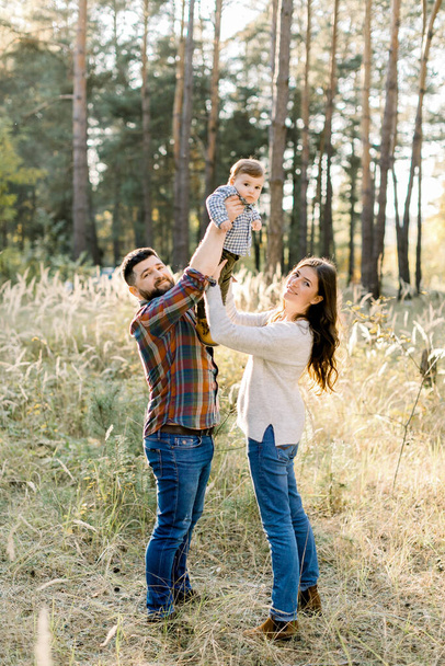 Outdoor family portrait of happy young parents, wearing stylish casual clothes, having fun and lifting up their little cute baby son, during walk in autumn forest at sunny day - Foto, Bild
