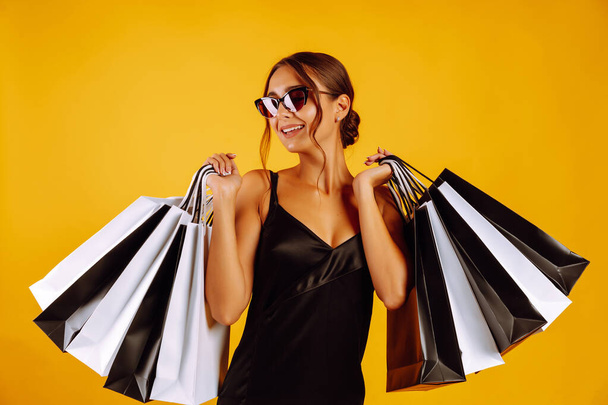 Fashion woman in black dress and sunglasses holding shopping bags. Young woman with black and white bags posing on yellow background. Purchases, black friday, discounts, sale concept. - Zdjęcie, obraz