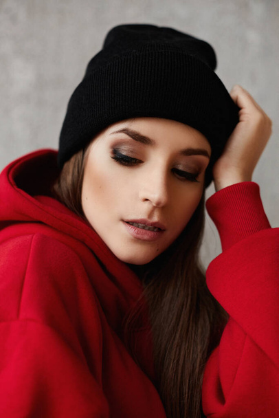 Portrait of a young woman with bright makeup in a black hat and red hoodie - Photo, Image