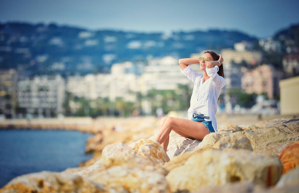 Mulher despreocupada sorrindo bonito em camisa branca e calções azuis está sentado em pedras perto do oceano. - Foto, Imagem