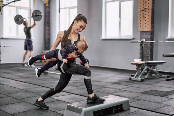 Beautiful fit woman holding her little son and doing lunges in the gym - 写真・画像