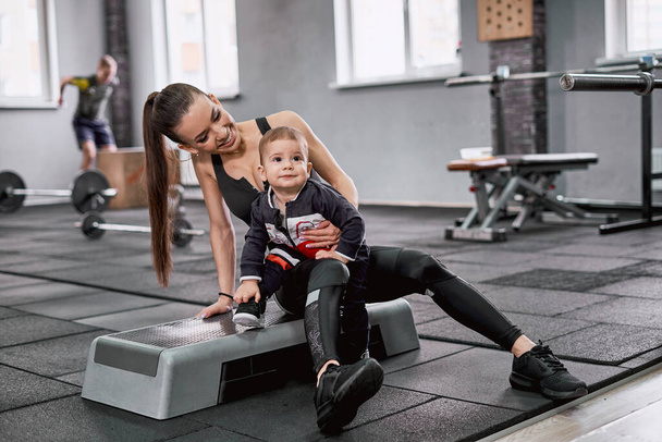 Portrait of a delighted woman with her little son sitting on a bench in the gym - Φωτογραφία, εικόνα
