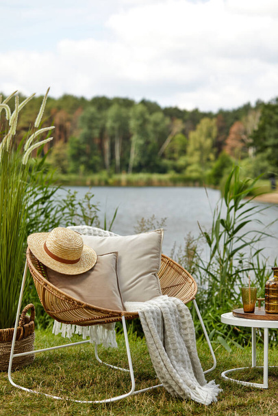 Stylish composition of outdoor garden on the lake with design rattan armchair, coffee table, plaid, pillows, drinks and elegant accessories. Summer chillout mood. - 写真・画像