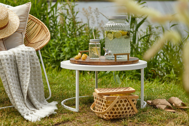 Stylish composition of outdoor garden on the lake with design rattan armchair, coffee table, plaid, pillows, drinks and elegant accessories. Summer chillout mood. - Photo, image