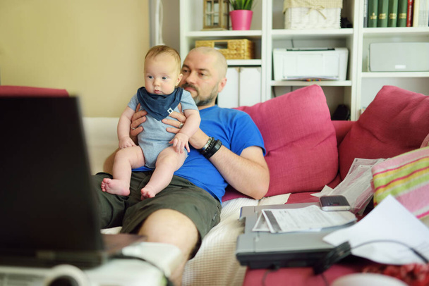 Young dad works remotely from home office with baby. Freelancer man holding his infant while using laptop. Workplace in living room. Working during quarantine. Staying at home. - Foto, Bild