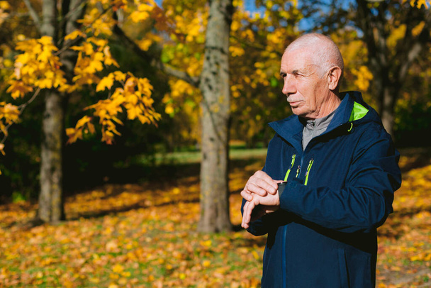 Old man using sport watches in autumn park. Senior athlete preparing for morning exercises outdoors. - Фото, зображення