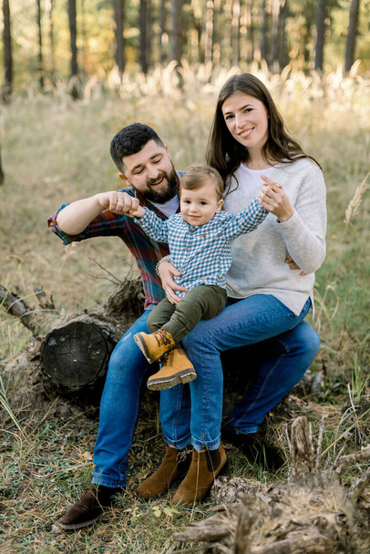 Outdoor lifestyle portrait of happy Caucasian family, father, mother and little baby boy, wearing stylish casual clothes, sitting on a log in pine autumn forest and smiling at camera - Foto, Bild