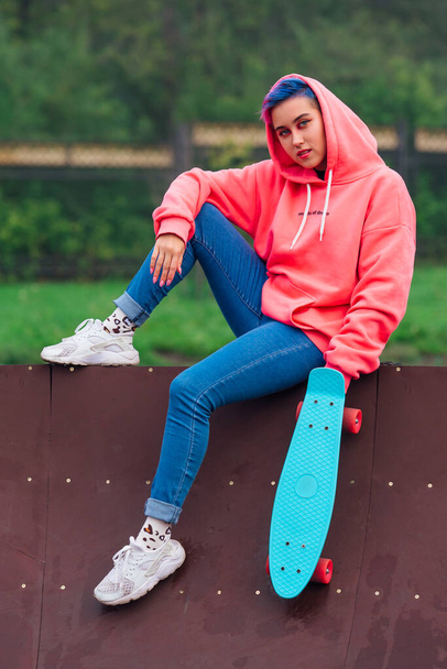 Young girl with short multicolored hair and nose piercing dressed in pink hoodie sits with skateboard in skatepark during morning fog. Portrait of a tomboy with plastic skateboard - Foto, immagini
