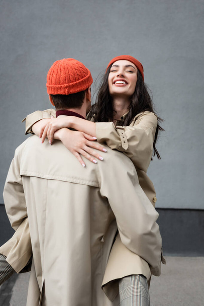 back view of man holding in arms joyful woman in beanie hat and trench coat  - Photo, Image