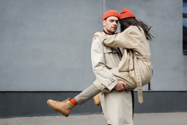 κομψό άνδρα κρατώντας στην αγκαλιά γυναίκα σε καπέλο σκούφο και καμπαρντίνα παλτό  - Φωτογραφία, εικόνα