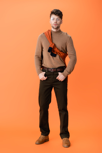 full length of stylish man in glasses standing with hands in pockets on orange  - Photo, image