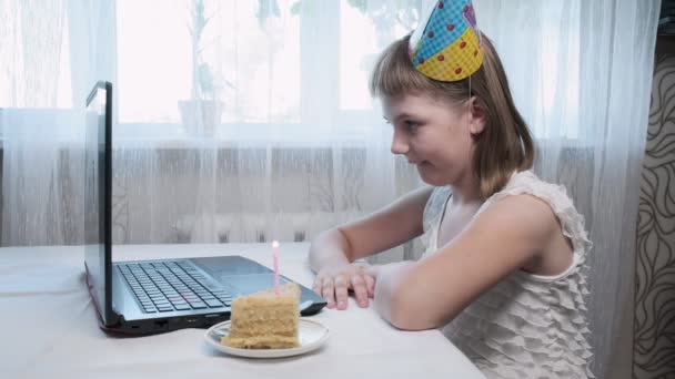 dívka sedí u stolu s notebookem, sfoukne svíčku na narozeninový dort - Záběry, video