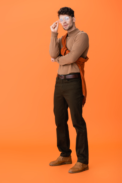 full length of trendy man in autumn outfit touching glasses on orange  - Photo, Image