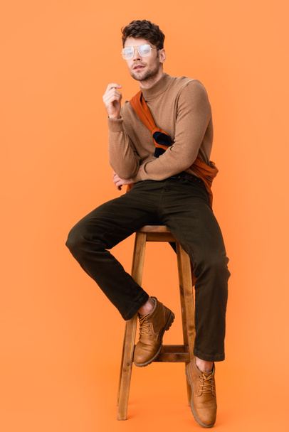 pensive man in autumn outfit and glasses sitting on wooden stool on orange  - Foto, immagini