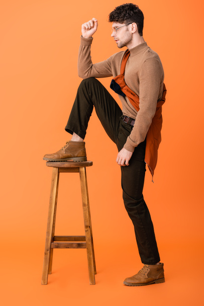 widok boczny stylowy mężczyzna w jesiennym stroju i okulary stojące z nogą na drewnianym stołku na pomarańczowym  - Zdjęcie, obraz