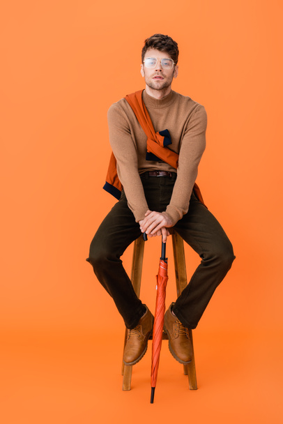 fashionable man in autumn outfit and glasses holding umbrella and sitting on wooden stool on orange  - Photo, Image