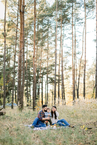 Lifestyle outdoor family portrait of young parents with little kid son, having fun together, while sitting on the blanket in beautiful autumn forest at sunny day - Foto, Imagem