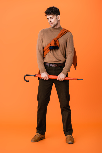 full length of stylish man in autumn outfit holding umbrella on orange  - 写真・画像
