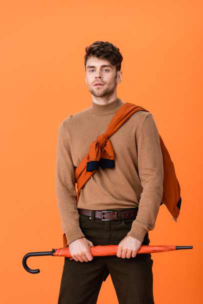 stylish man in autumn outfit holding umbrella while looking at camera isolated on orange  - Photo, Image