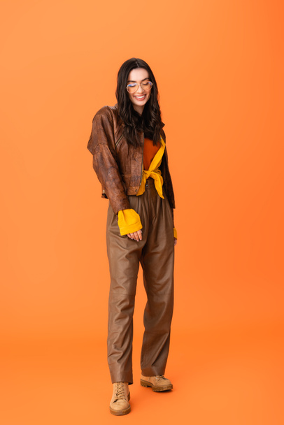 full length of young woman in autumn outfit and glasses standing on orange  - 写真・画像