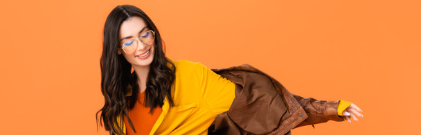panoramatický záběr stylové ženy v brýlích a kožené bundě izolované na oranžové  - Fotografie, Obrázek