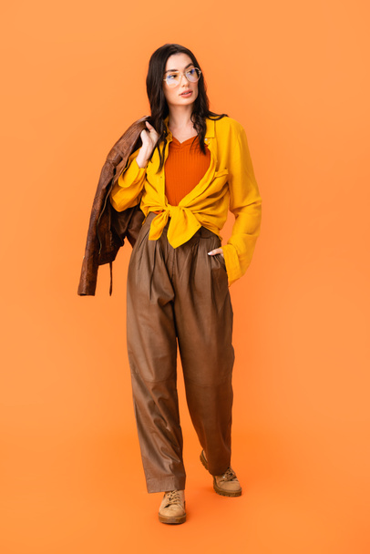full length of young woman in autumn outfit holding leather jacket and standing with hand in pocket on orange  - Foto, Imagem