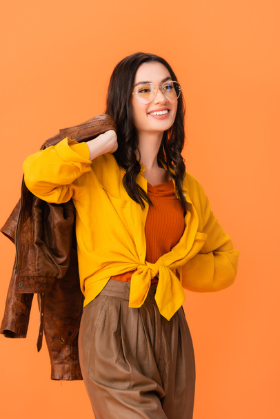 trendy woman in glasses and autumn outfit holding leather jacket while standing with hand on hip isolated on orange  - Fotografie, Obrázek