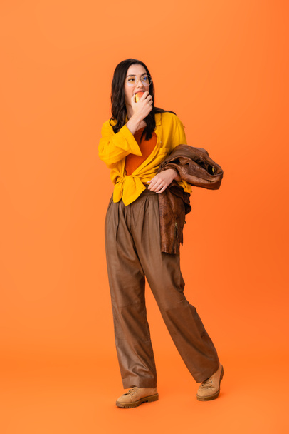 πλήρες μήκος της μοντέρνας γυναίκας σε γυαλιά και φθινοπωρινό ντύσιμο κρατώντας δερμάτινο μπουφάν και τρώγοντας ώριμο μήλο σε πορτοκαλί - Φωτογραφία, εικόνα