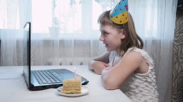meisje zit aan tafel met laptop computer, blaast aangestoken kaars op verjaardag taart - Video