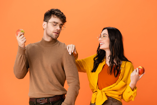 couple élégant dans des lunettes et tenue d'automne tenant des pommes tout en se regardant isolé sur orange - Photo, image