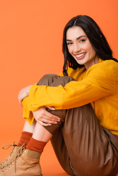joyful young woman in autumn outfit and boots looking at camera while sitting isolated on orange  - Photo, Image