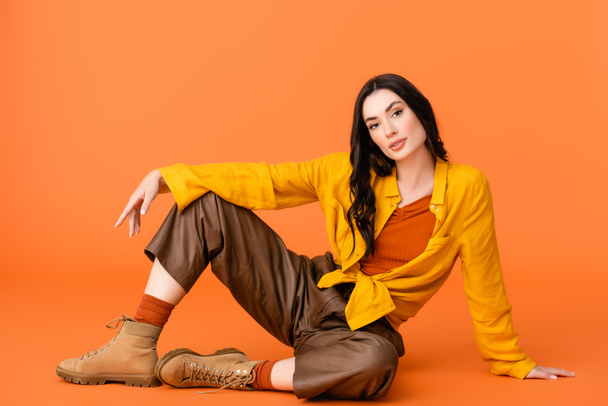 модная молодая женщина в осеннем наряде и сапогах, смотрящая в камеру, сидя на оранжевом  - Фото, изображение