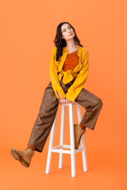 full length of young woman in autumn outfit and boots sitting on white stool on orange  - Фото, зображення