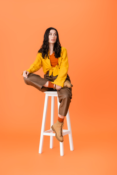 femme à la mode en tenue d'automne assis sur tabouret blanc et regardant loin sur orange  - Photo, image