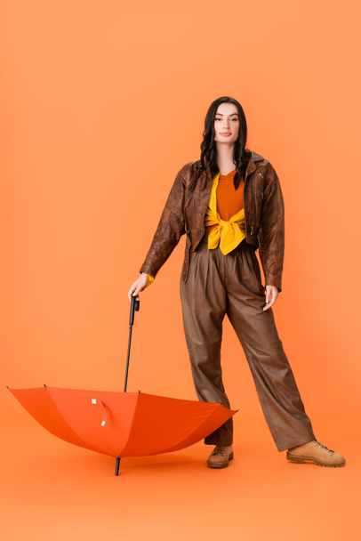 pełna długość modnej kobiety w jesiennym stroju i buty stojące z parasolem na pomarańczowym - Zdjęcie, obraz