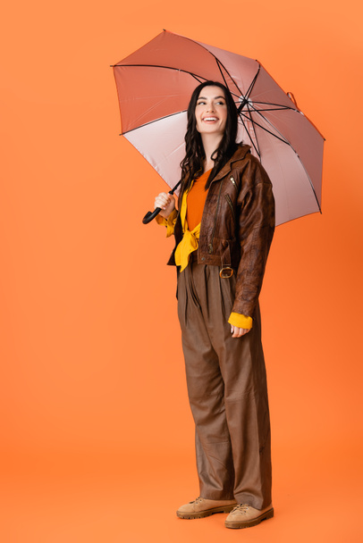 full length of joyful woman in autumn outfit and boots standing with umbrella on orange - Photo, Image
