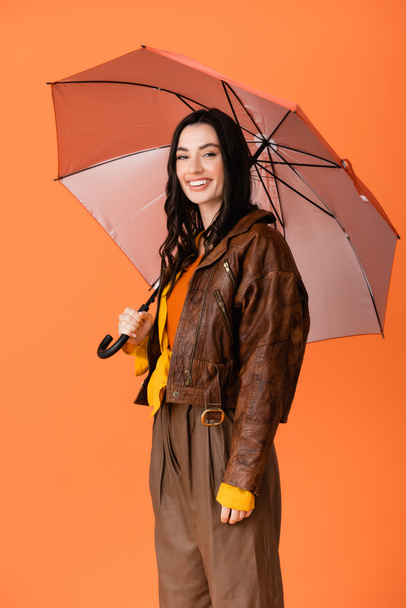 young woman in autumn outfit standing under umbrella isolated on orange - Zdjęcie, obraz