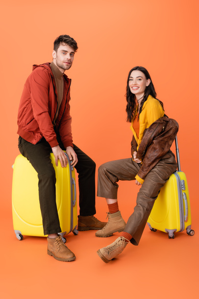 joyful and trendy couple in autumn outfit sitting on yellow baggage on orange  - Photo, Image