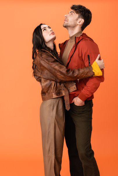 brunette woman hugging stylish man standing with hand in pocket and looking up on orange - 写真・画像