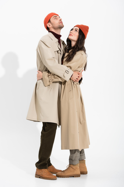 full length of trendy couple in beanie hats and trench coats hugging and looking up on white  - Фото, изображение